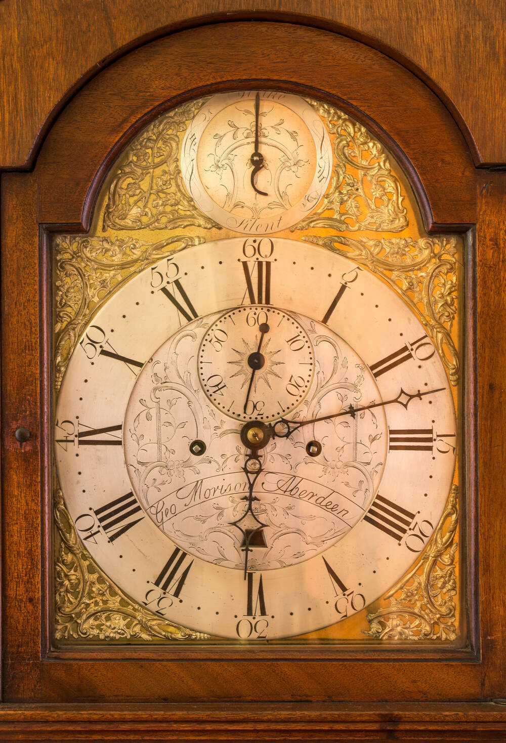 A close-up of a wooden clock’s face, engraved with the maker’s name. It has roman numerals around a white large dial, with a smaller dial above.