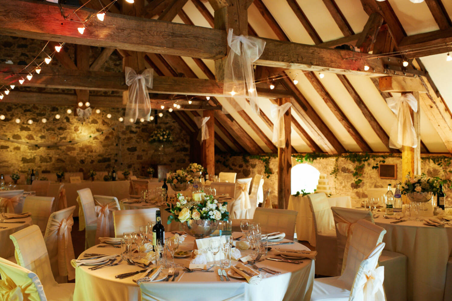 Inside a stone barn with wooden rafters, strung with fairy lights. Round tables have been set up for a wedding reception.