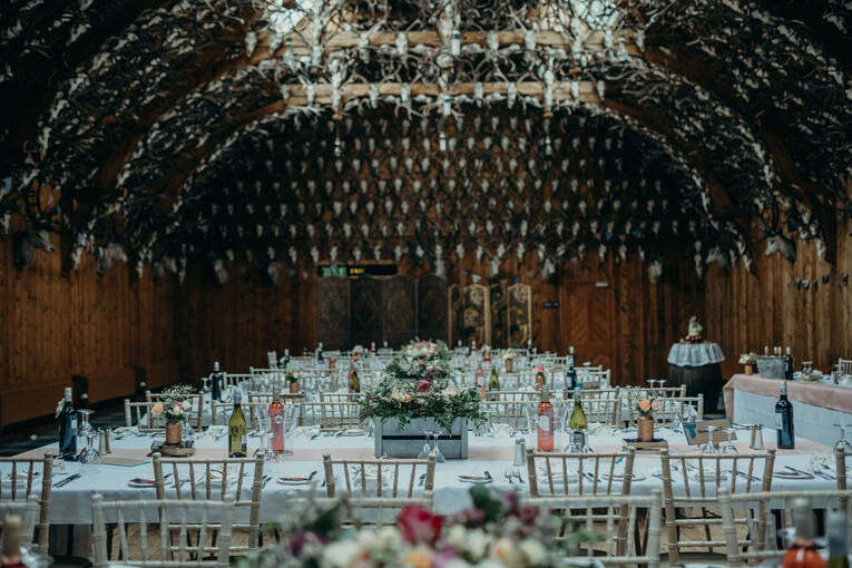 A room with an arched ceiling, with the upper walls covered in stags' heads, set up with several tables for a wedding reception.