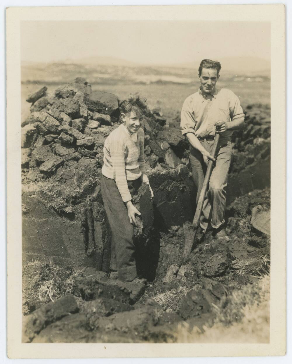 Black and white photograph of two mean cutting peat in a landscape.