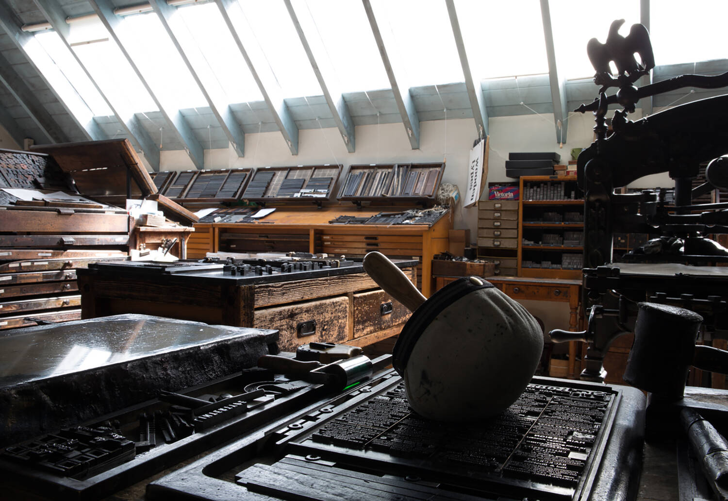 A view of the caseroom at Robert Smail's Printing Works. A cloth hammer lies on type of a case of set type in the foreground. In the background are many shelves filled with pieces of type.
