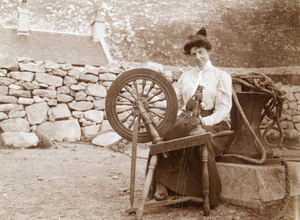 Black and white image of a woman sitting at a spinning wheel. There is a drystone wall behind her.