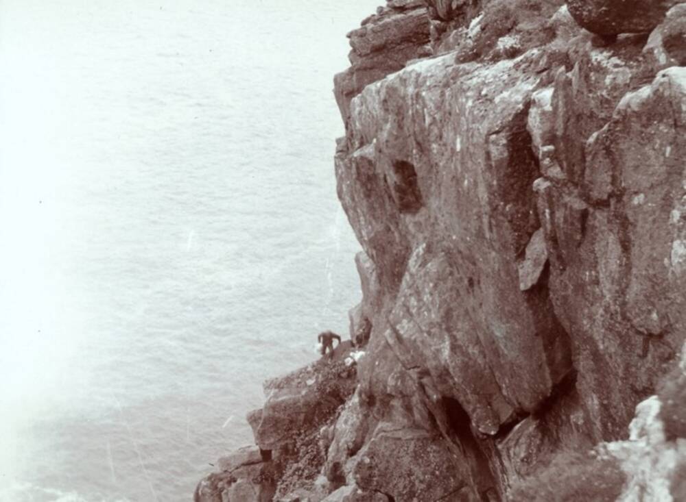 Black and white image of a man precariously perched on the side of a high sea cliff.