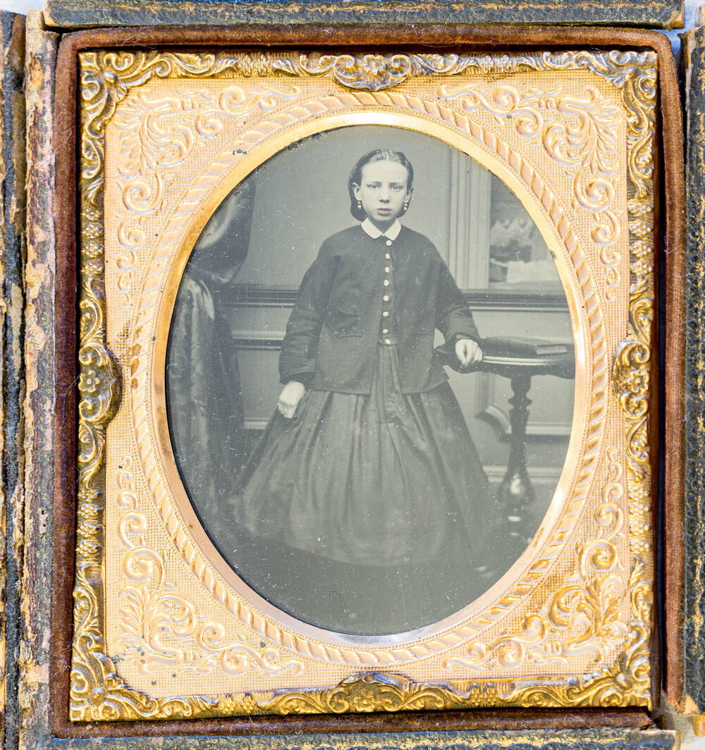 An oval black and white photo of a girl in a dark dress holding onto a table. The photo is in a gilded frame.