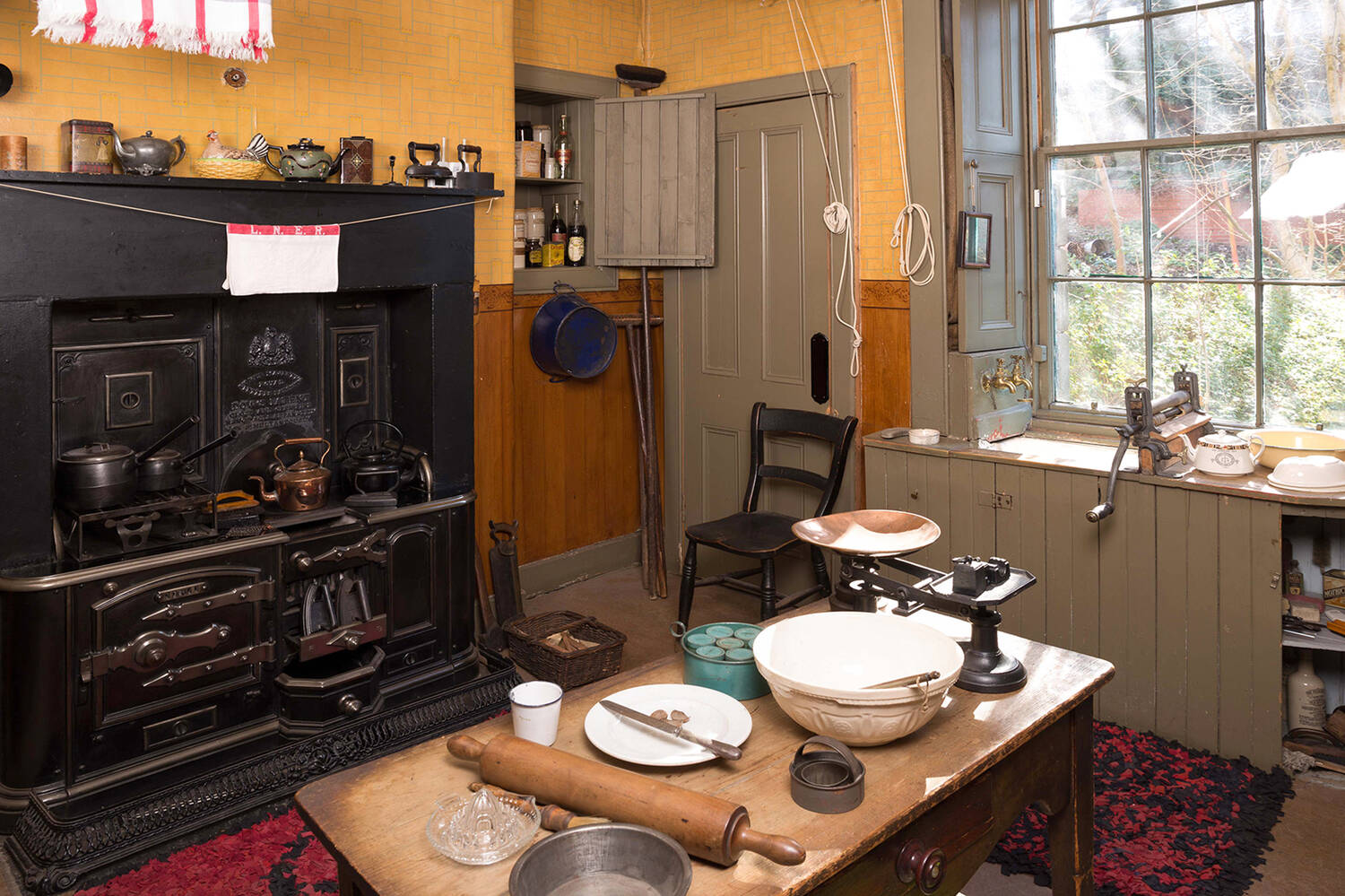 A view of a kitchen in a tenement flat, displayed as it may have looked in the mid-20th century. A large wooden table stands in the middle of the room, with mixing bowls, weighing scales and a rolling pin laid out on top. A black cooking range stands at the side, with cloths hanging from a washing line suspended in front. A sink with wooden cupboards beneath stands in front of a large sash window.