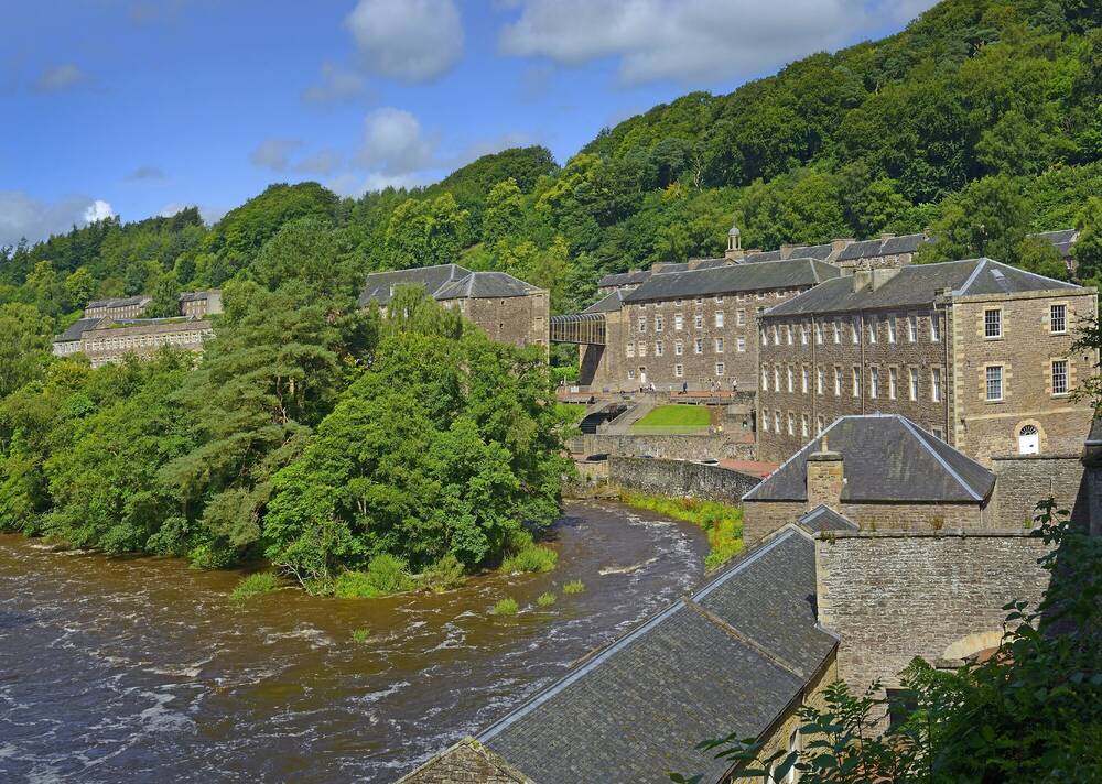 A cluster of stone mill buildings and houses on the banks of a river, surrounded by trees.
